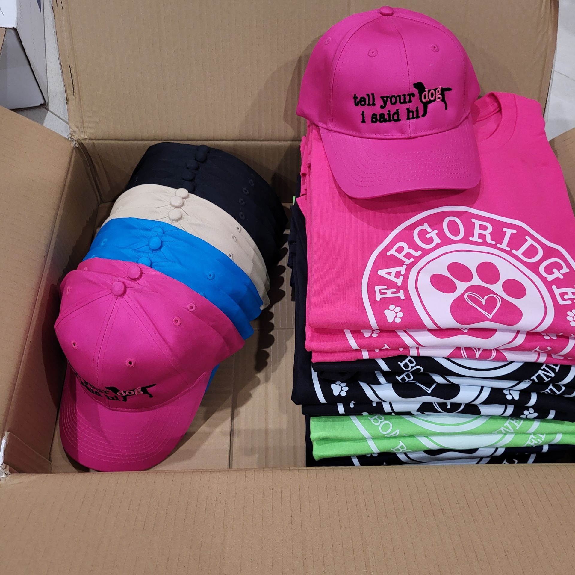Box of pink and black hats and t-shirts with various logos and colors.