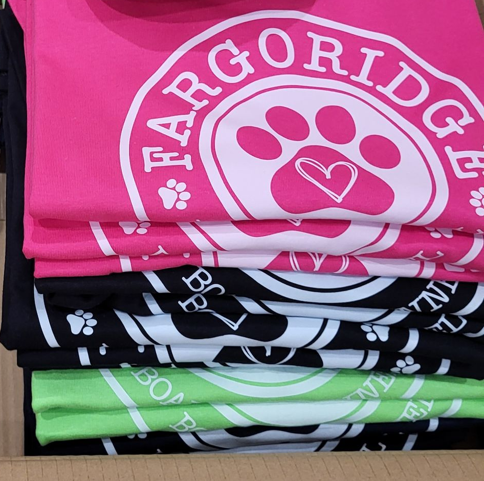 Stacks of folded t-shirts with a paw print logo, in pink, white, black, and green.