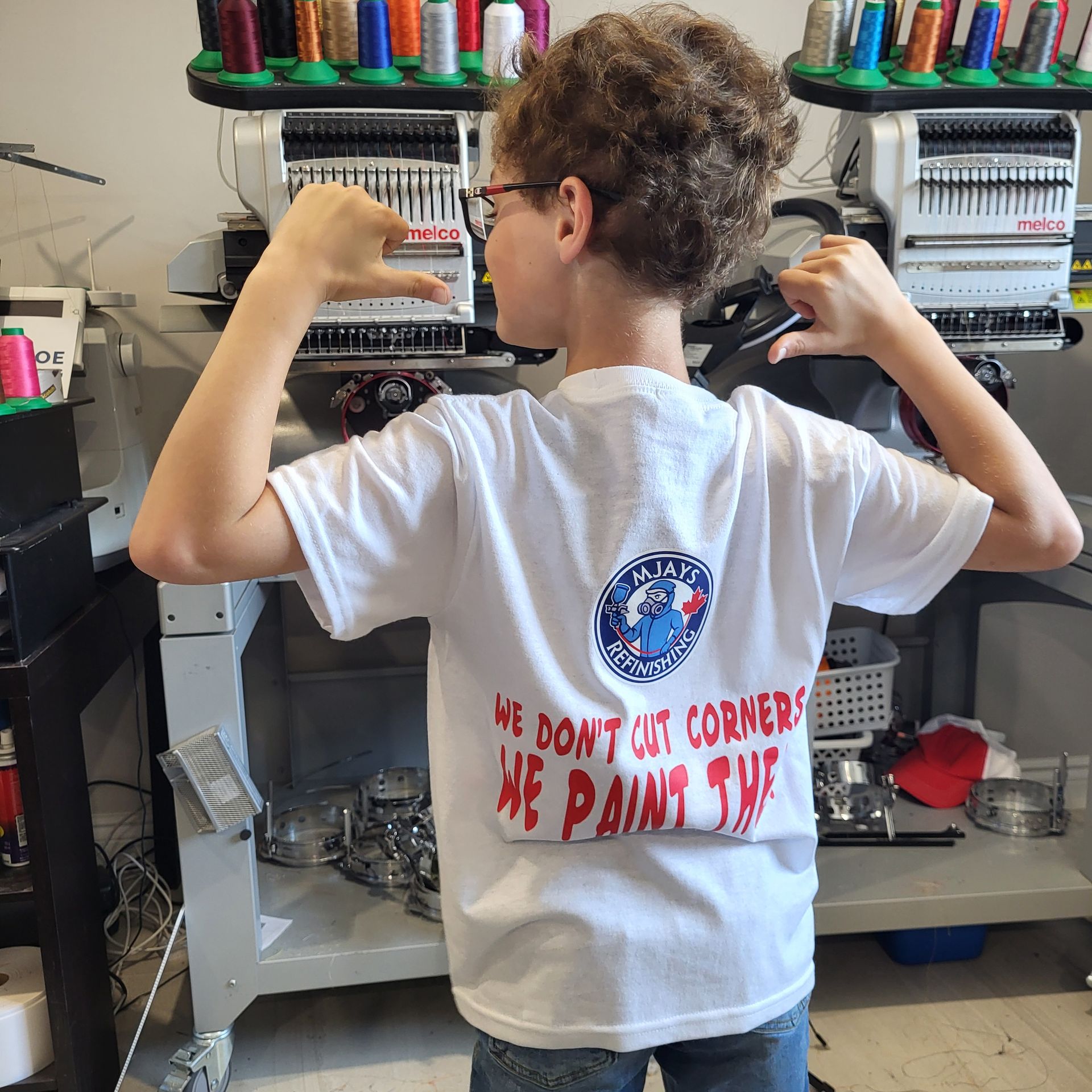 A boy wearing a t-shirt with logo, standing in front of embroidery machines.