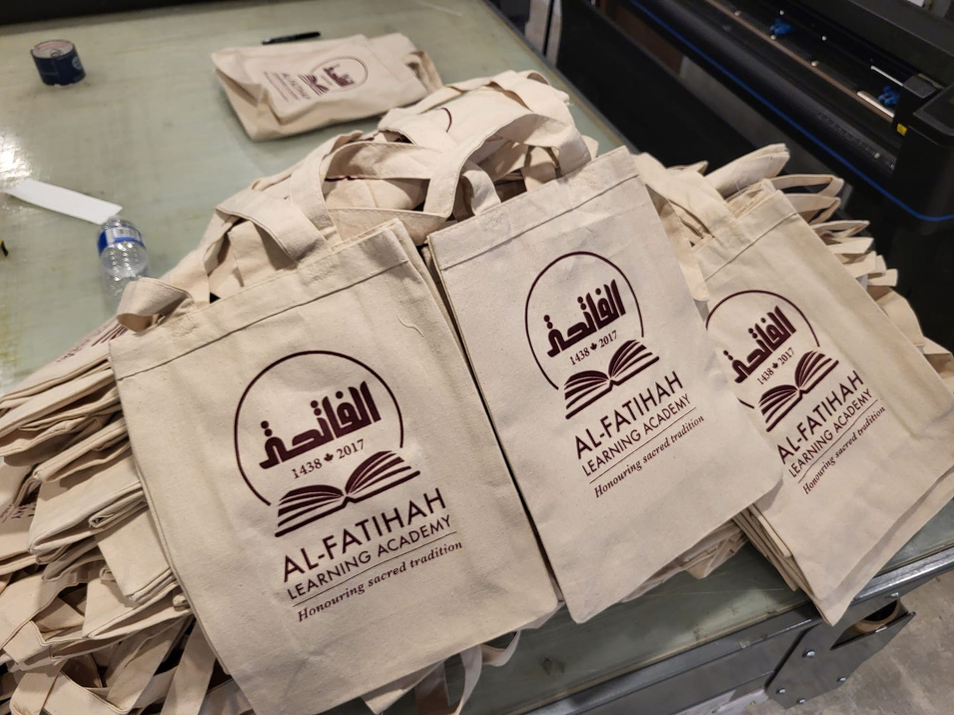 Beige tote bags printed with a maroon logo and text, stacked on a table.