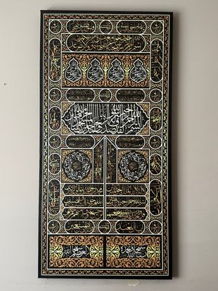 Black and gold Islamic calligraphy art on a black framed canvas, against a light tan wall.