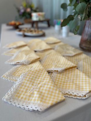 Yellow gingham napkins with lace trim on a light-colored table; cake and flowers in the background.