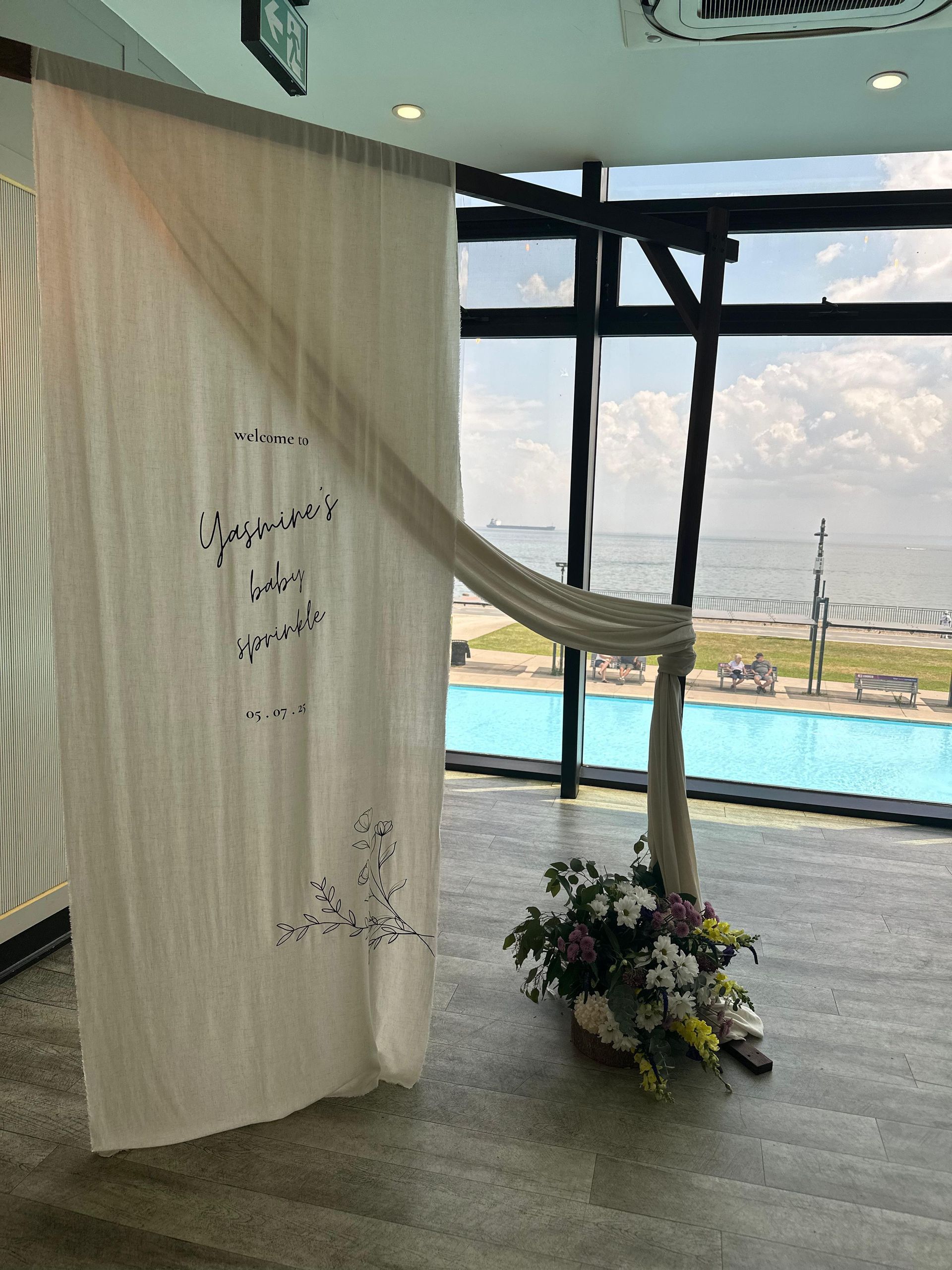 White curtain, draped on black beams, by a window overlooking a pool and ocean, with floral arrangement at the base.