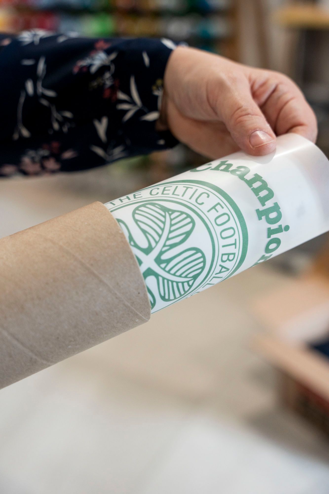Hand holding a poster with a green Celtic Football Club logo rolled in a cardboard tube.