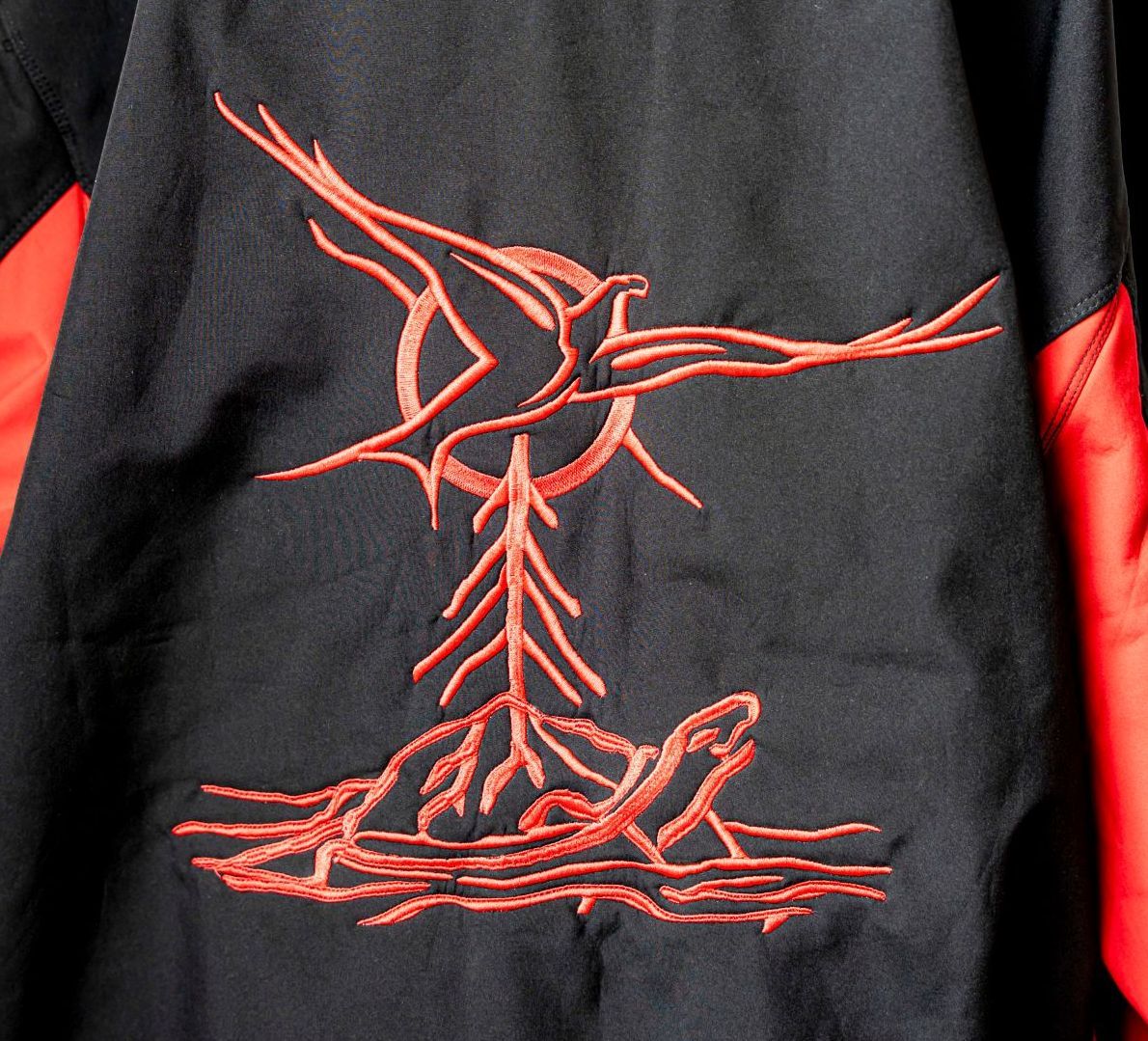 Red embroidered design on black jacket: bird above a tree-like shape over a turtle.
