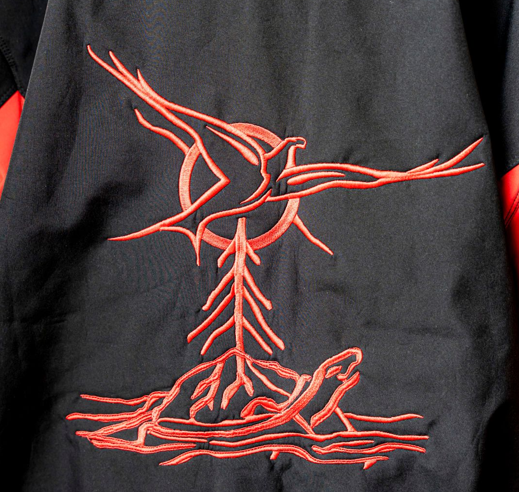 Red embroidered design on a black jacket. Features a bird above a tree-like form and a turtle.