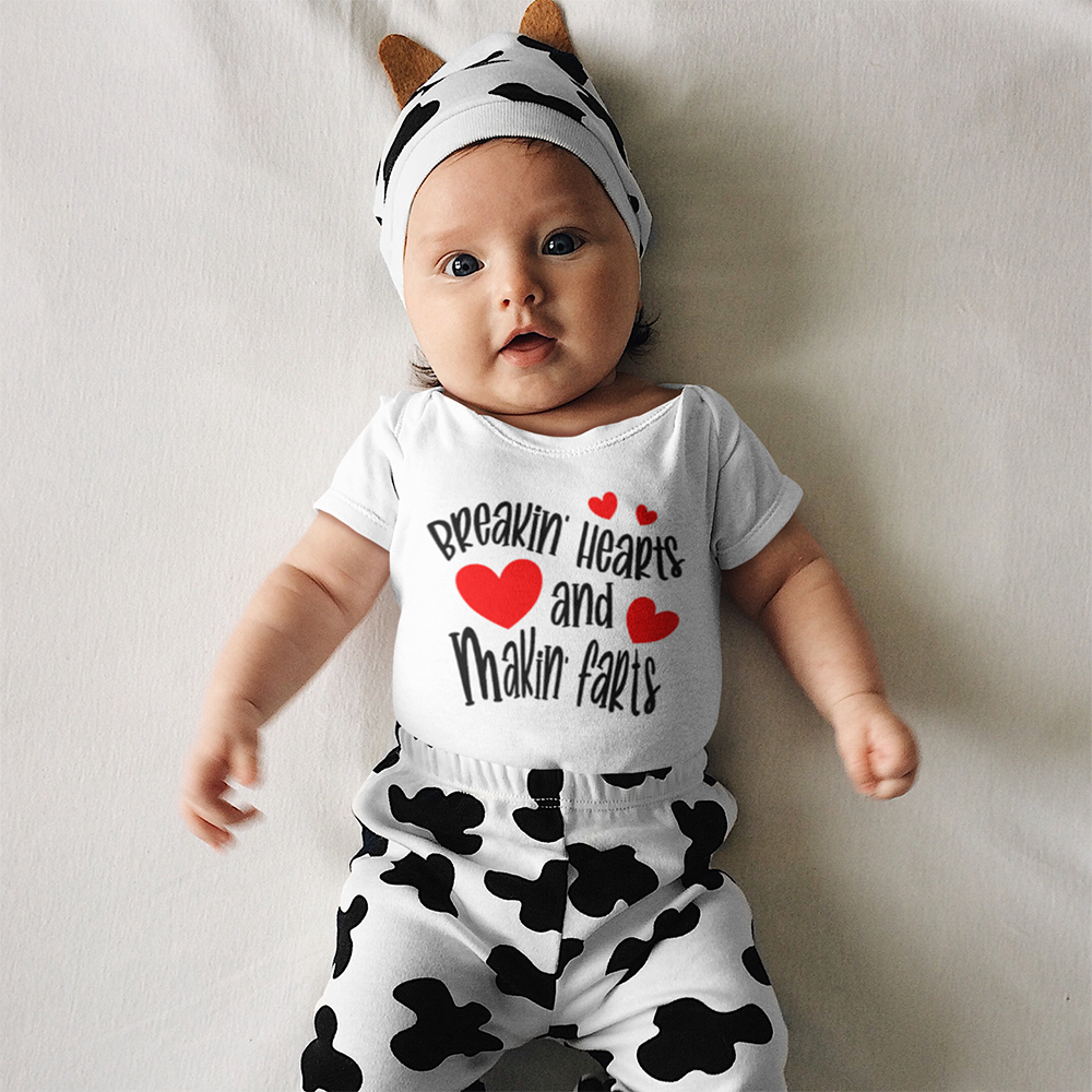 Baby in cow-print outfit, wearing hat with ears, laying on white surface. Outfit reads