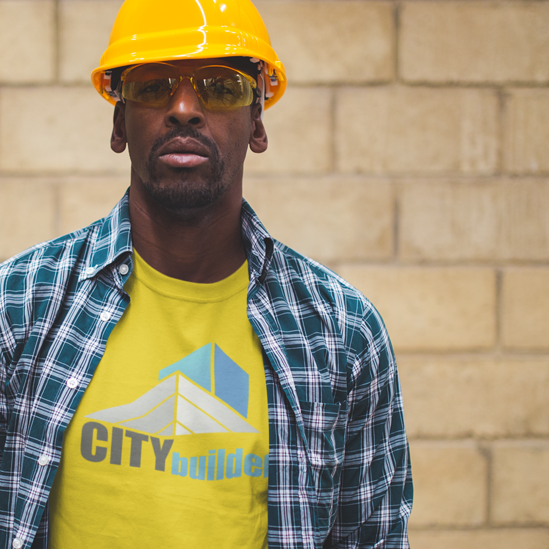 Construction worker wearing yellow helmet, glasses, and plaid shirt; City Builder logo on shirt; brick wall.