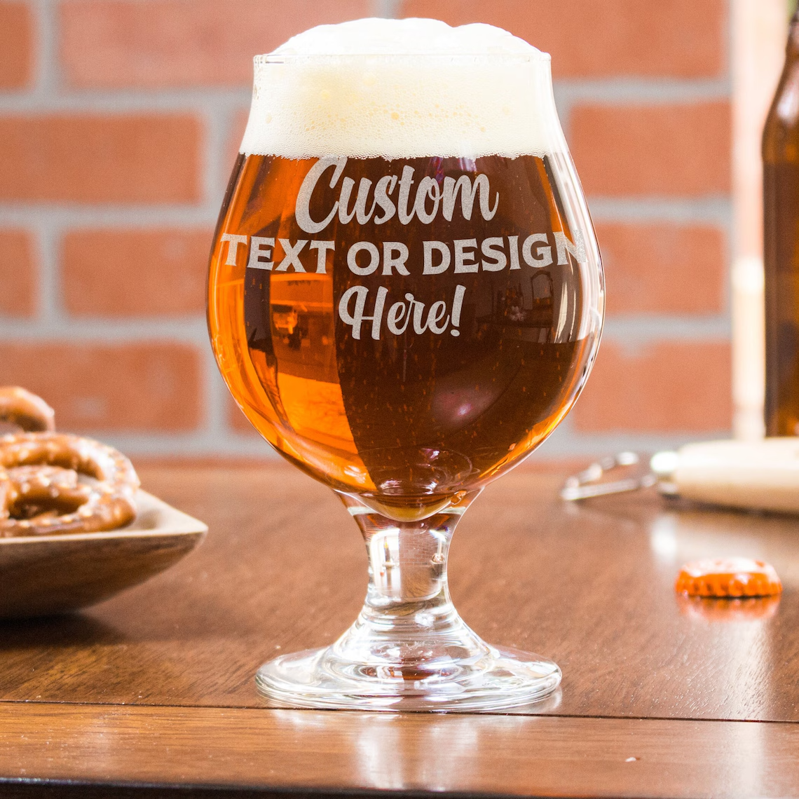 Beer glass with foamy head, customized text, on a wooden table with pretzels and a bottle.