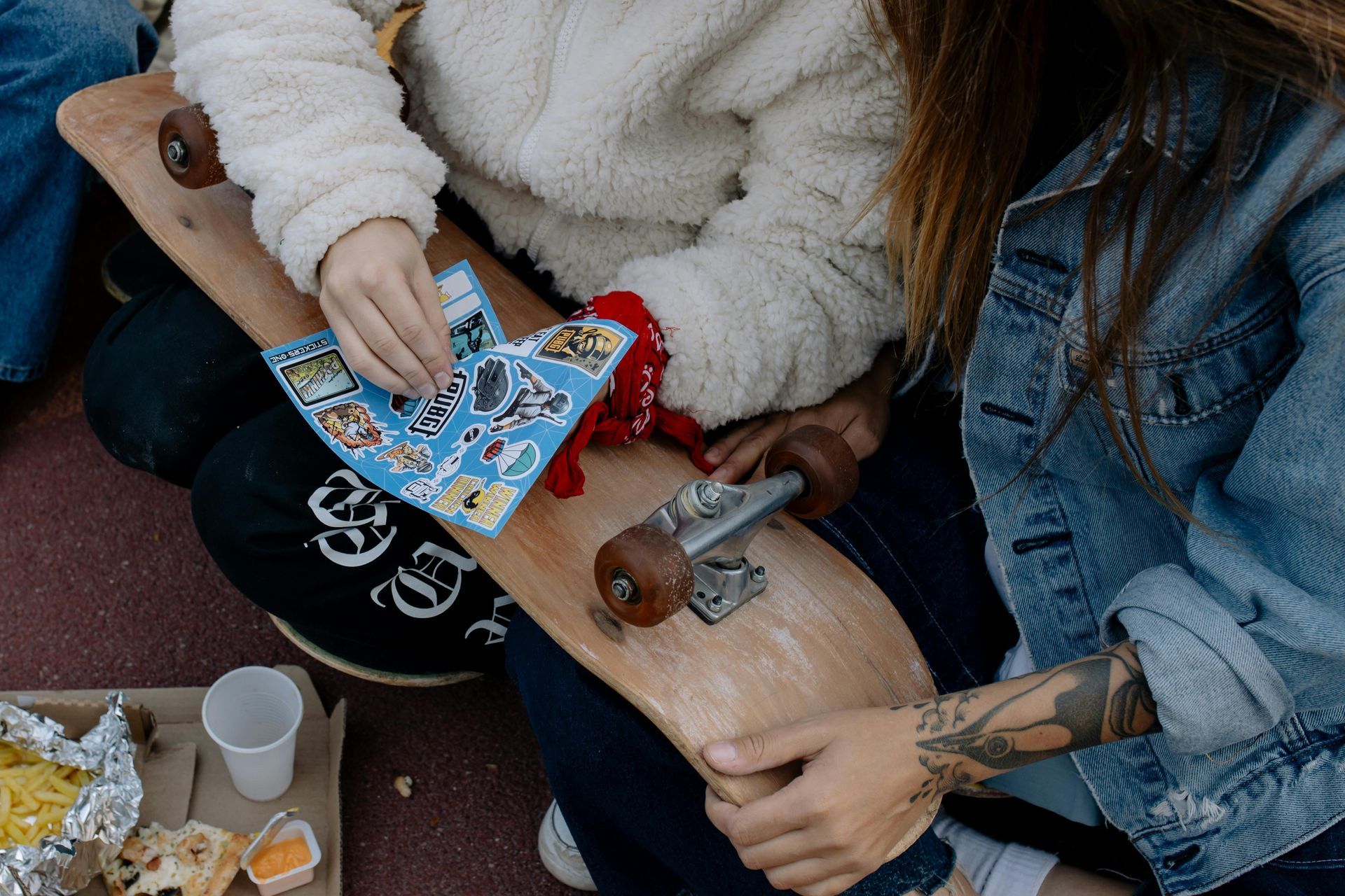 Two people applying stickers to a skateboard outdoors. One wears a denim jacket and has tattoos.