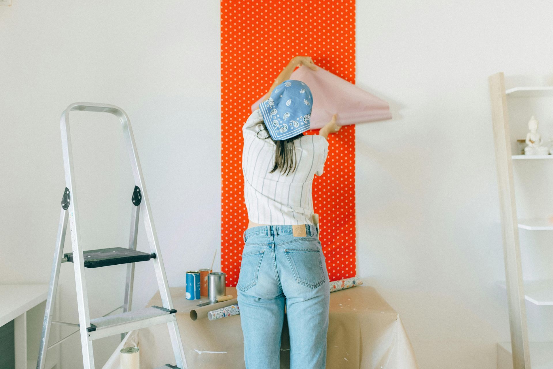Person wallpapering a red dotted wall, standing on a stepstool in a room with a ladder and shelf.