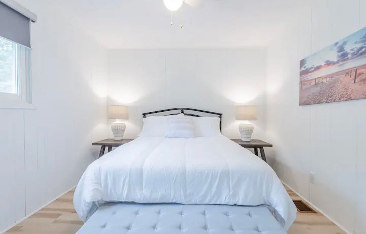 A bedroom with a bed , ottoman , lamps and a painting on the wall.