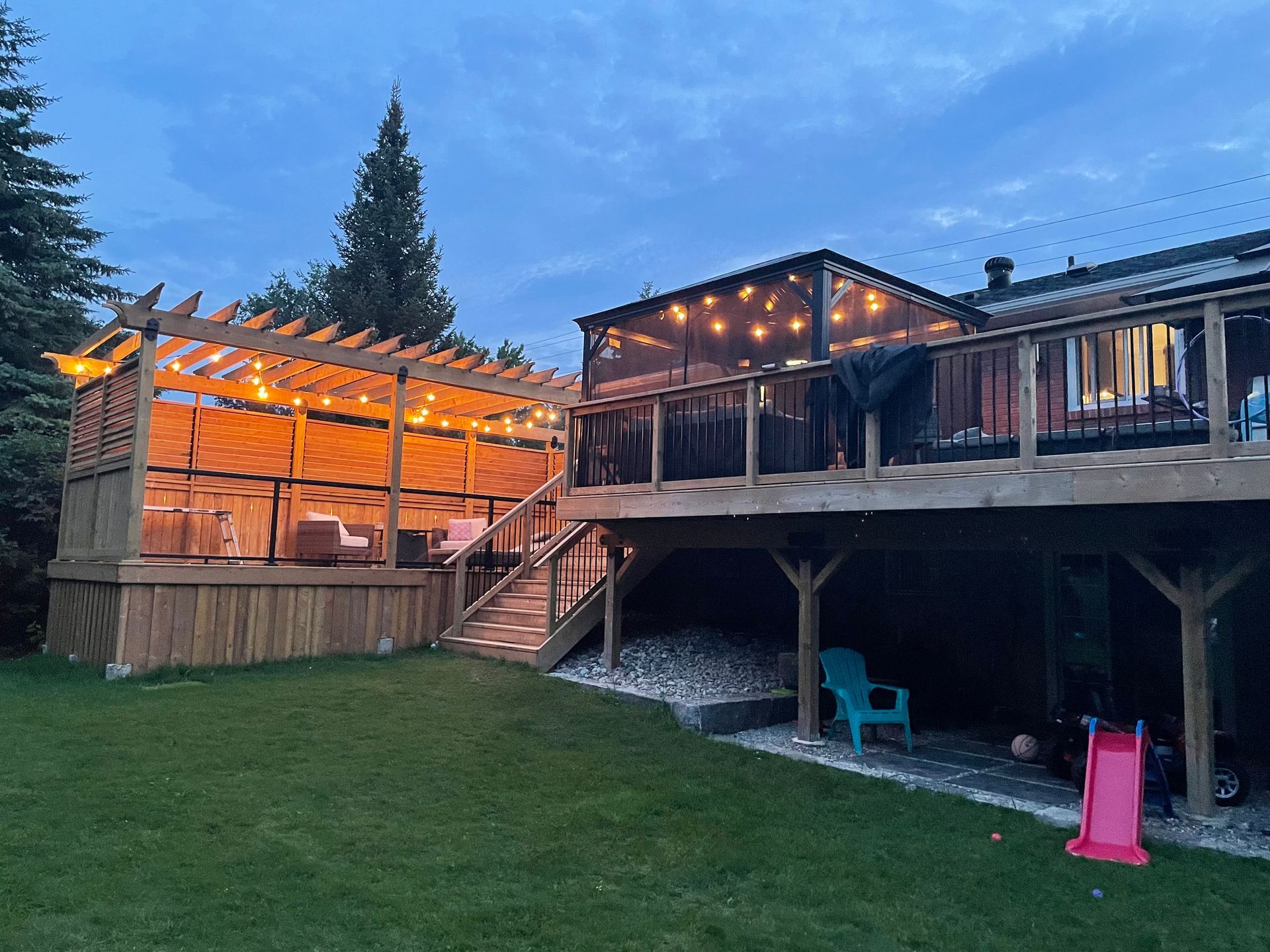 A large wooden deck with a pergola and lights on it is in the backyard of a house.