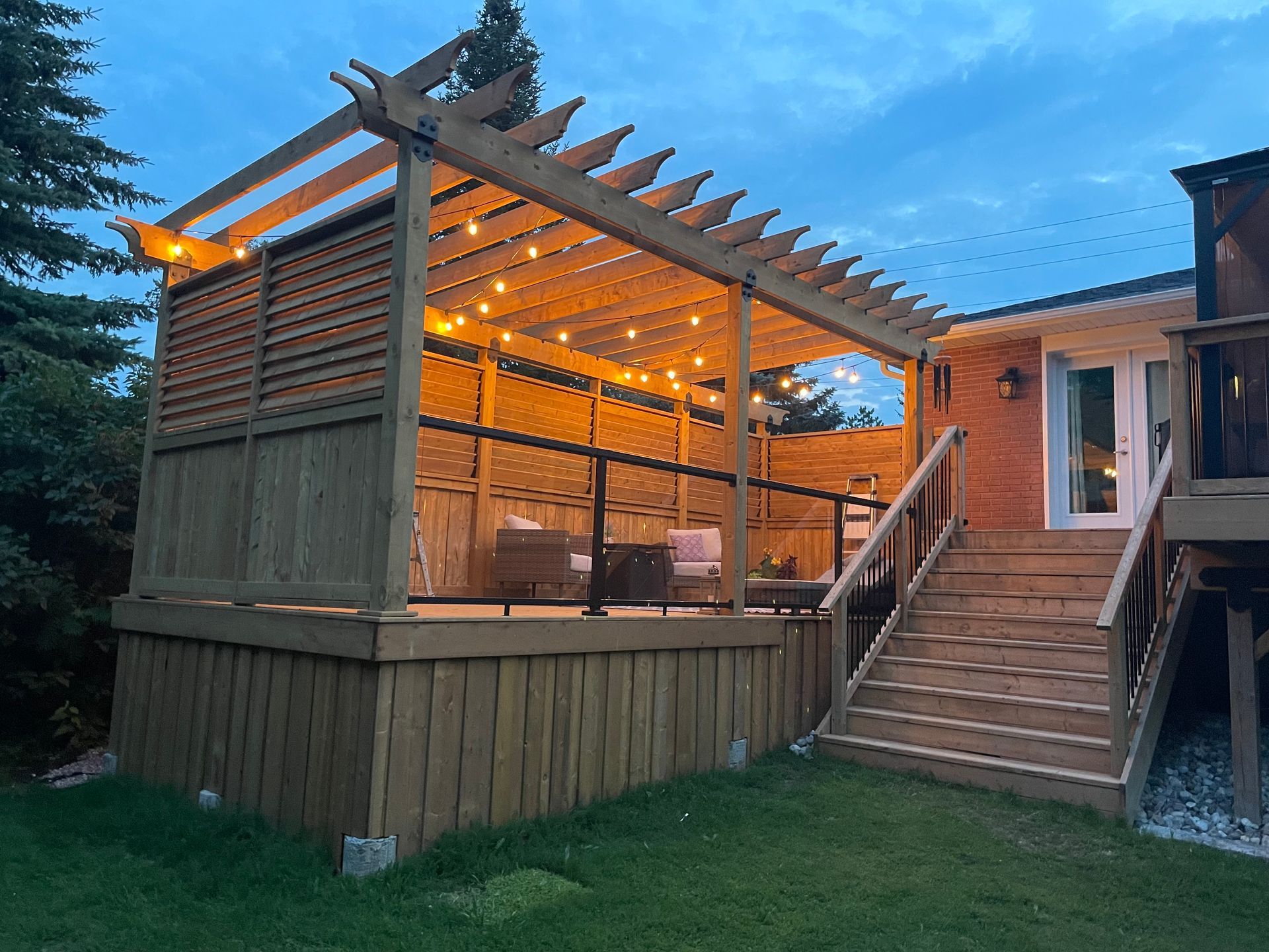 A wooden deck with a pergola and stairs is lit up at night.