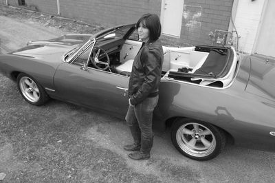 A woman in a leather jacket is standing next to a convertible car