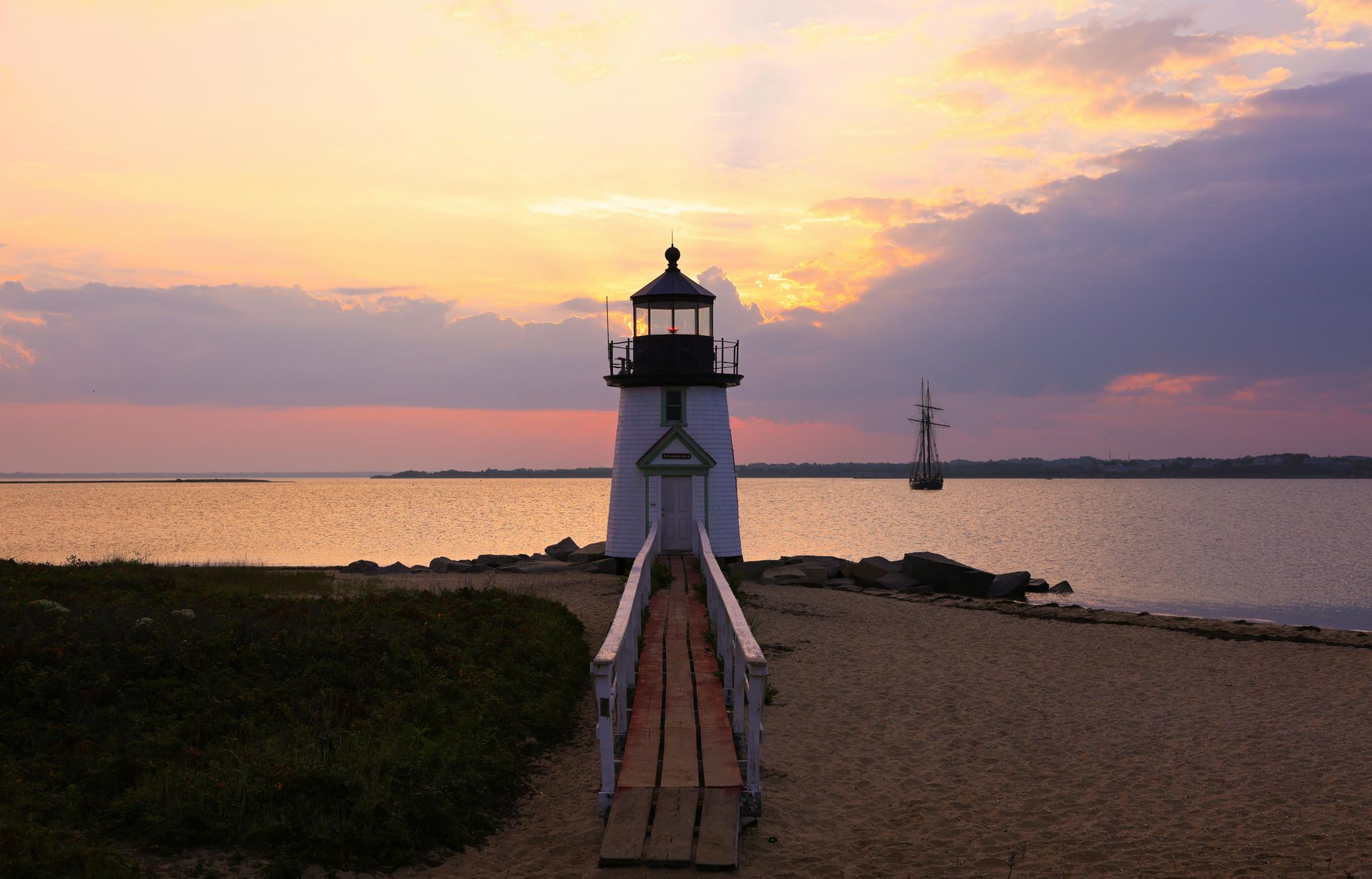 A white lighthouse at sunset, with a wooden walkway leading to it and a tall ship sailing in the distance.