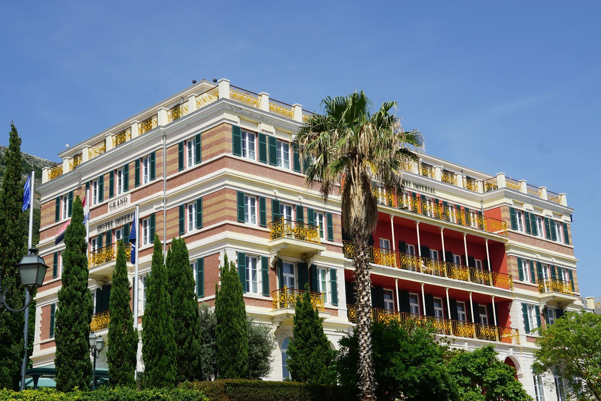 Hotel building with red and tan facade, green shutters, and palm tree.