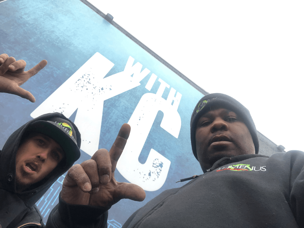 Two men pose in front of a sign that says kc