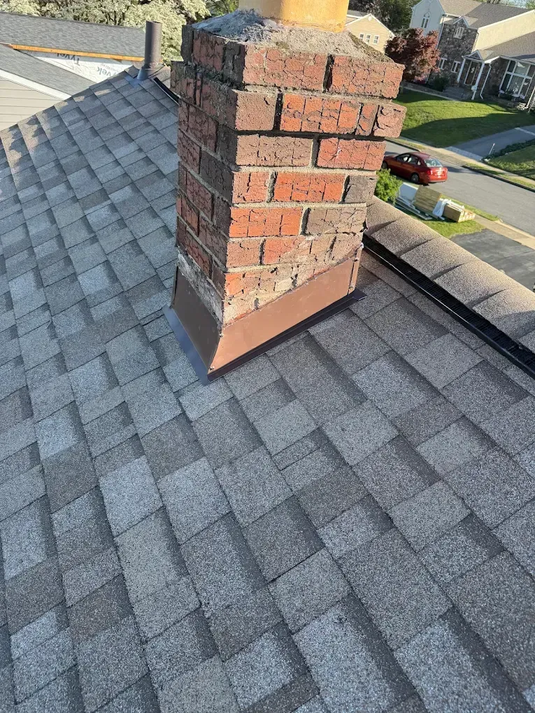 Brick chimney on a gray shingled roof with nearby homes in the background