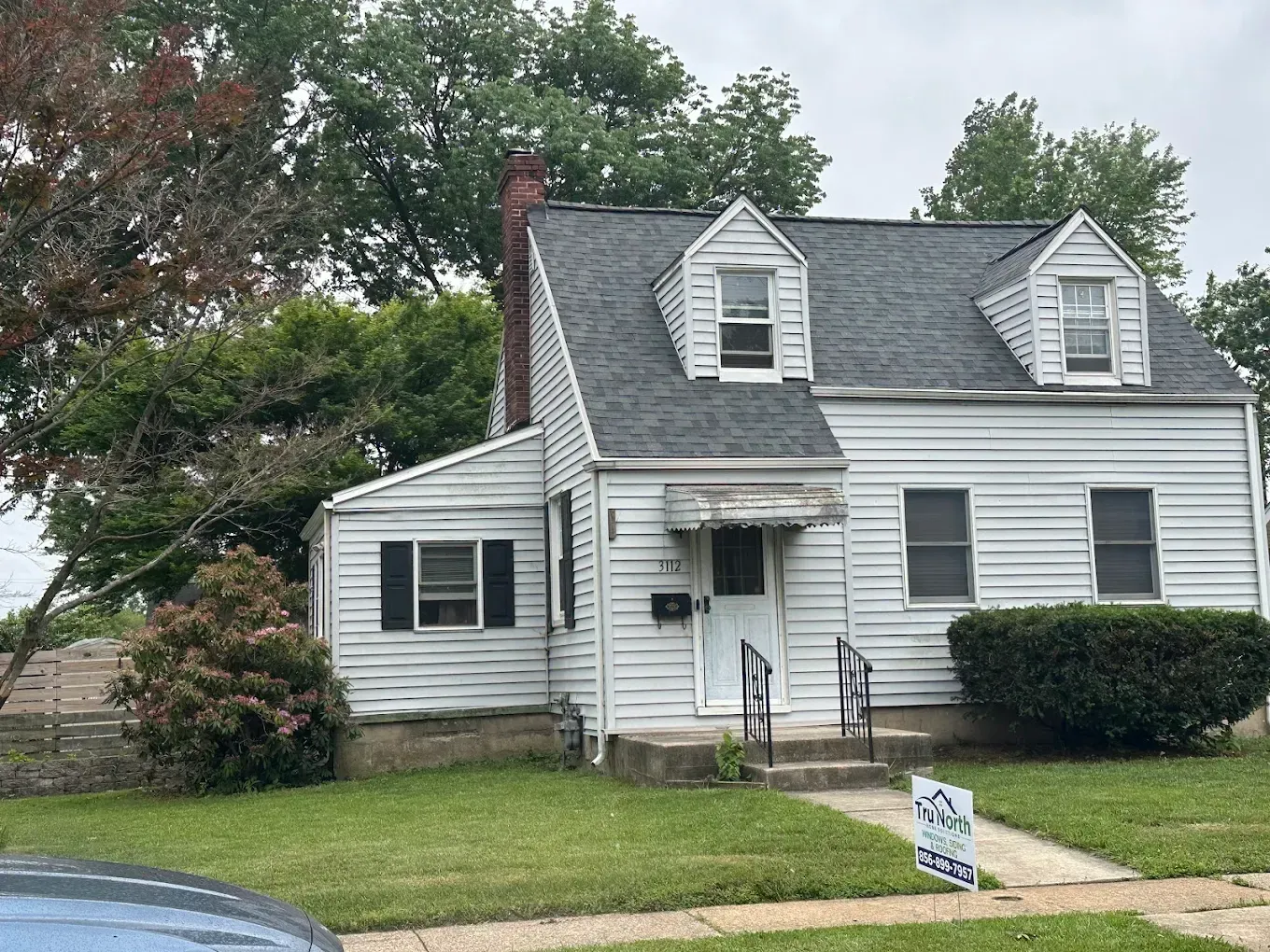 Small white house with dormer windows, front porch, and lawn, viewed from the street with a sign in front.