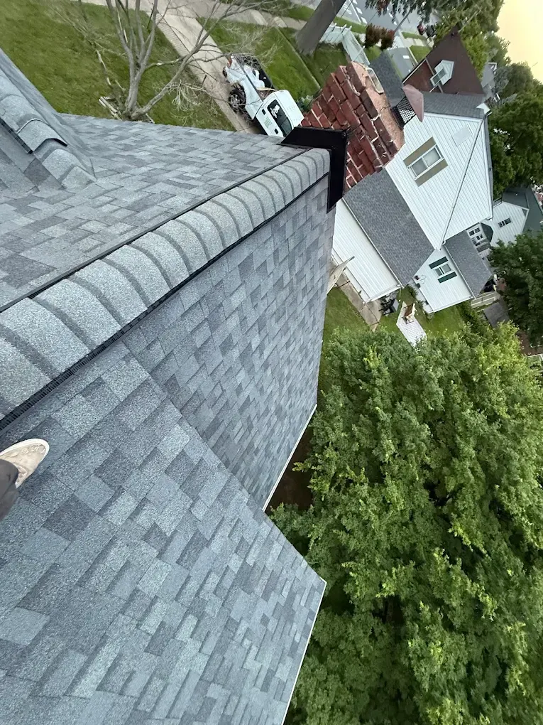 View from a steep gray shingle roof above a white house and green trees in a suburban neighborhood.