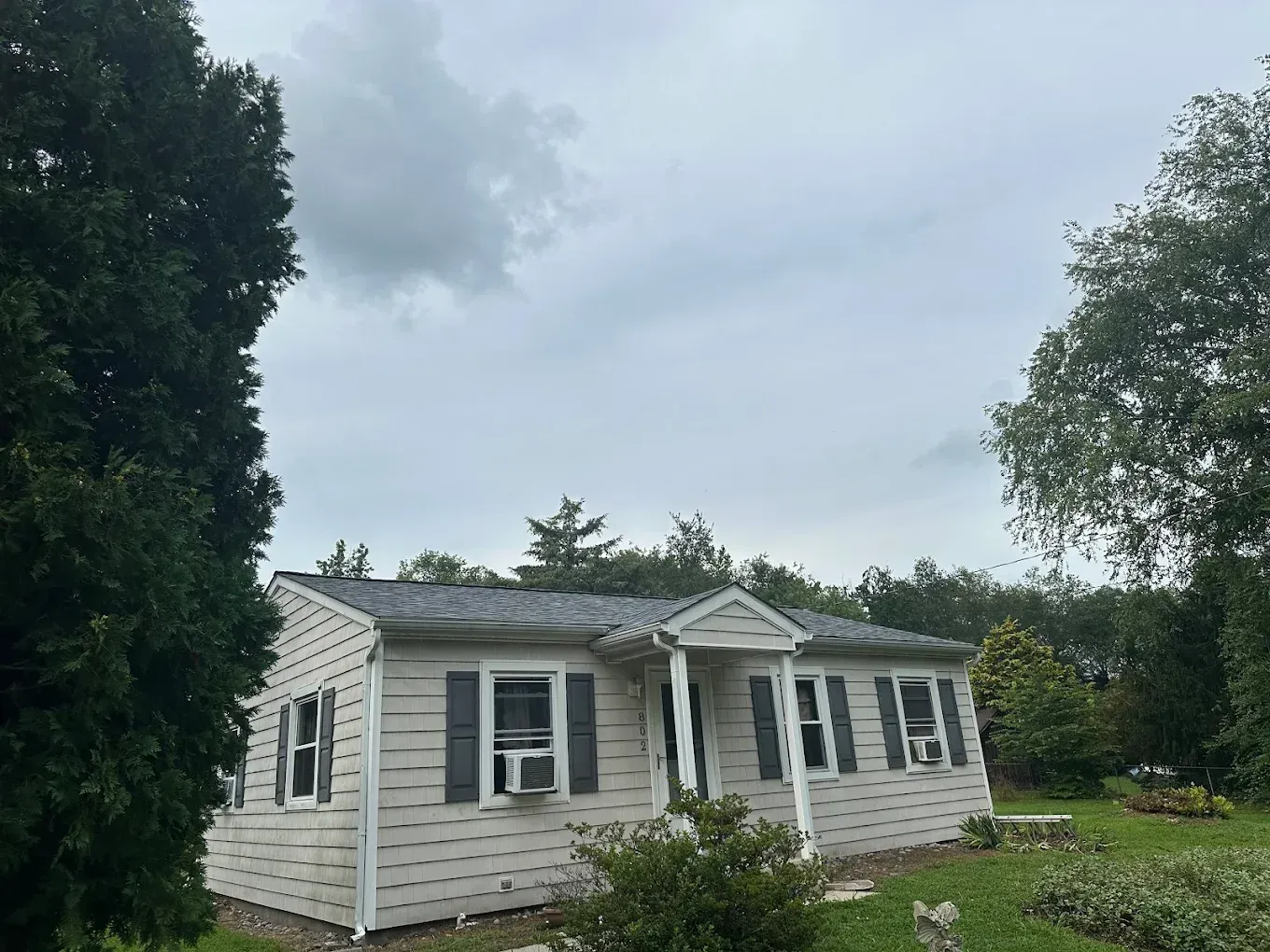 Small white cottage with dark shutters and a front porch, surrounded by trees under a cloudy sky