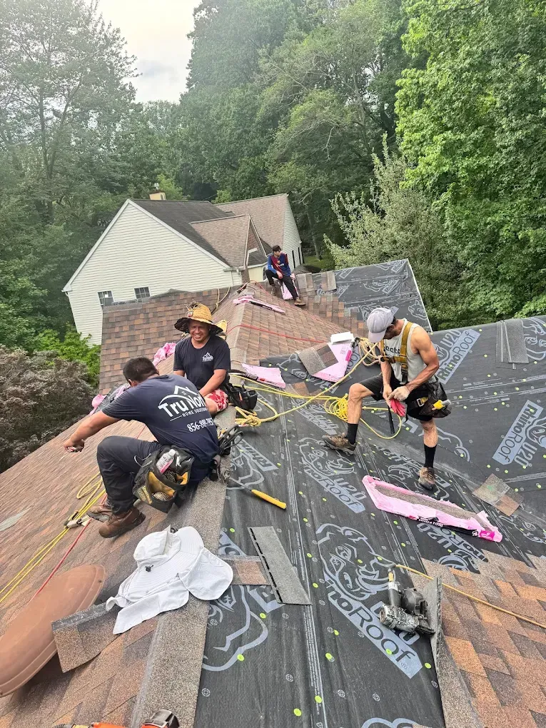 Roofers repairing a damaged roof in a wooded area, using tools and laying shingles near a house.