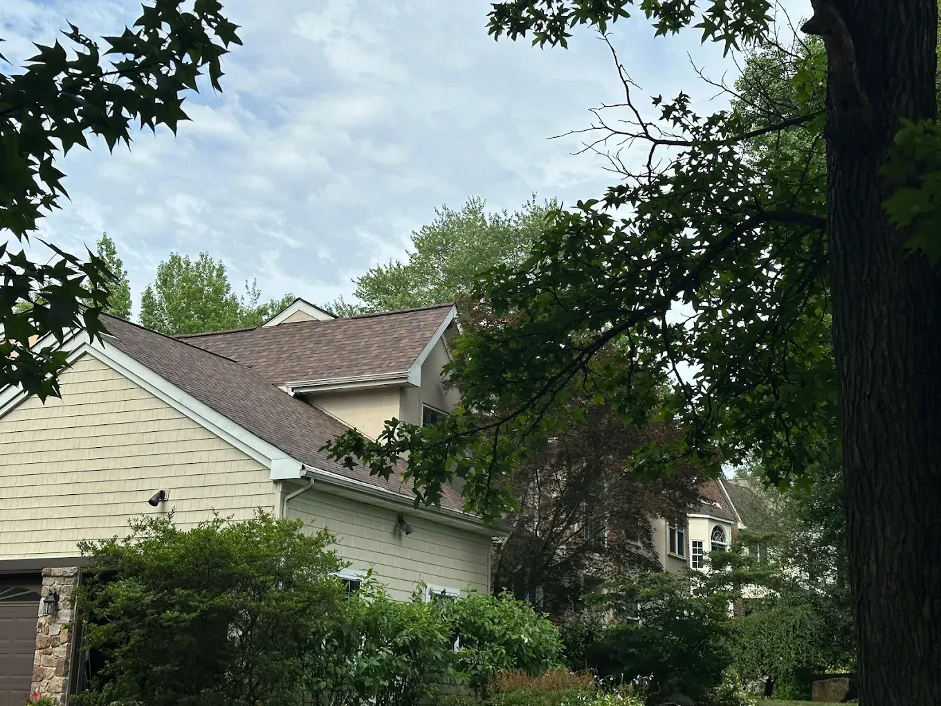 Suburban houses with beige siding and brown roofs, partially obscured by leafy trees and bushes