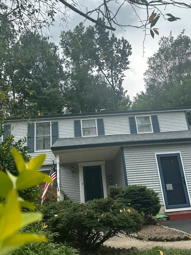 Two-story gray house with blue doors and white trim, partly hidden by trees and shrubs.