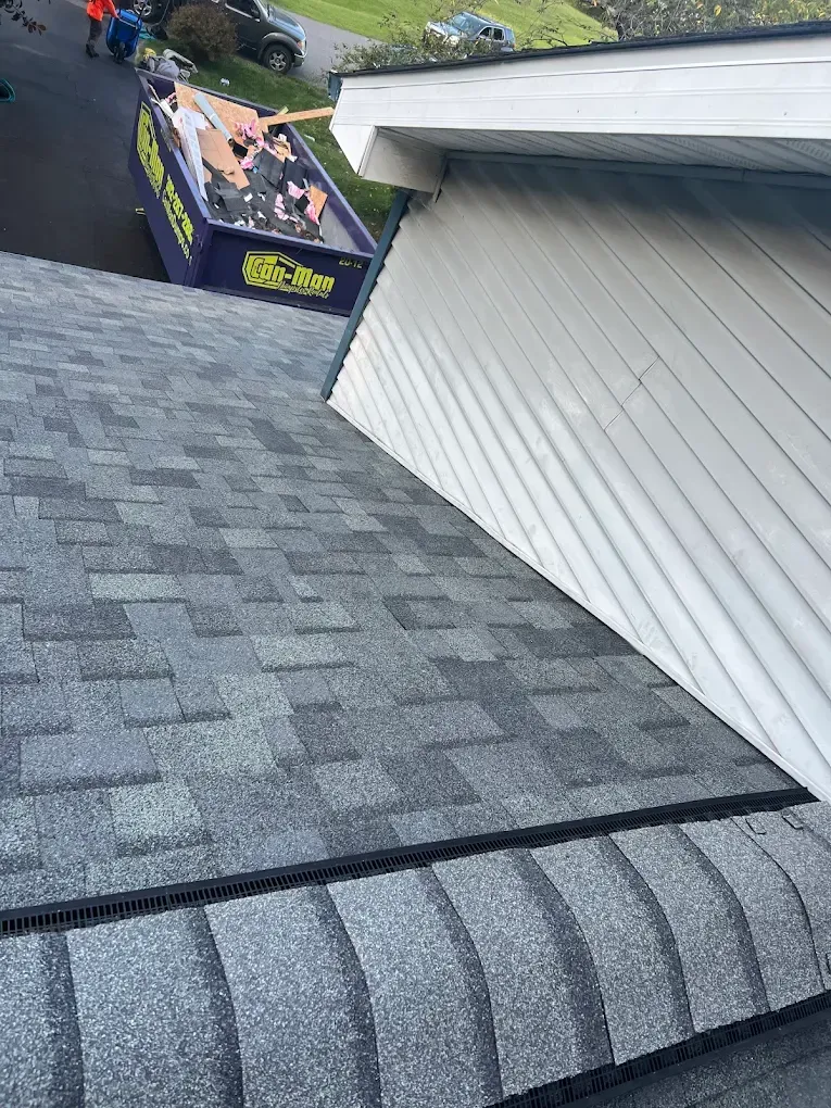 Roof shingles beside white siding, with a dumpster and wet street below.