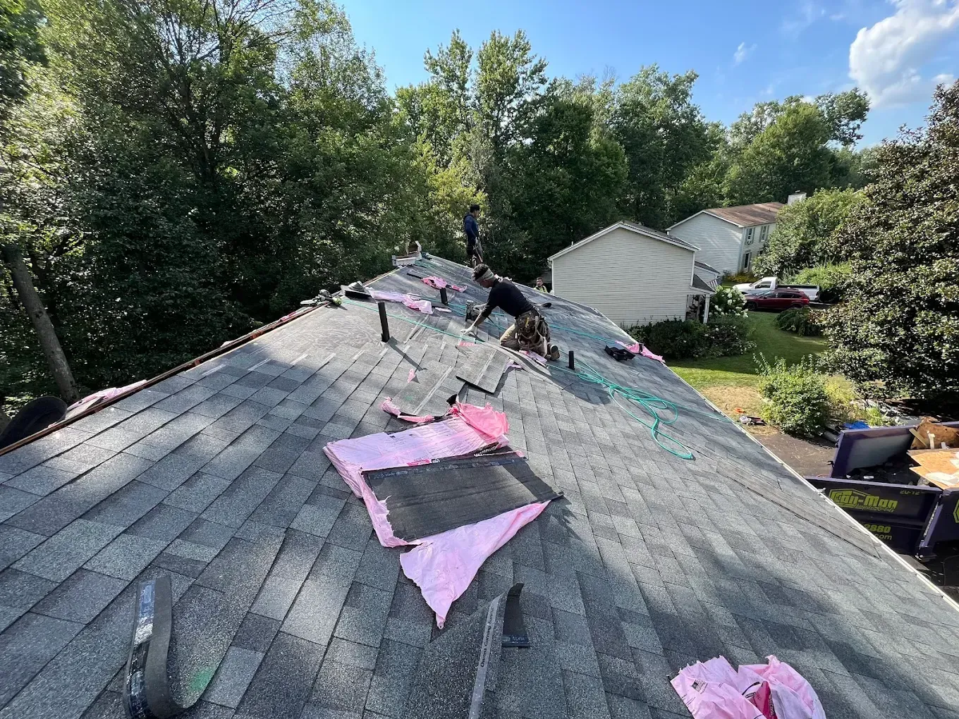 Damaged shingled roof with pink patches and exposed underlayment near a house and trees