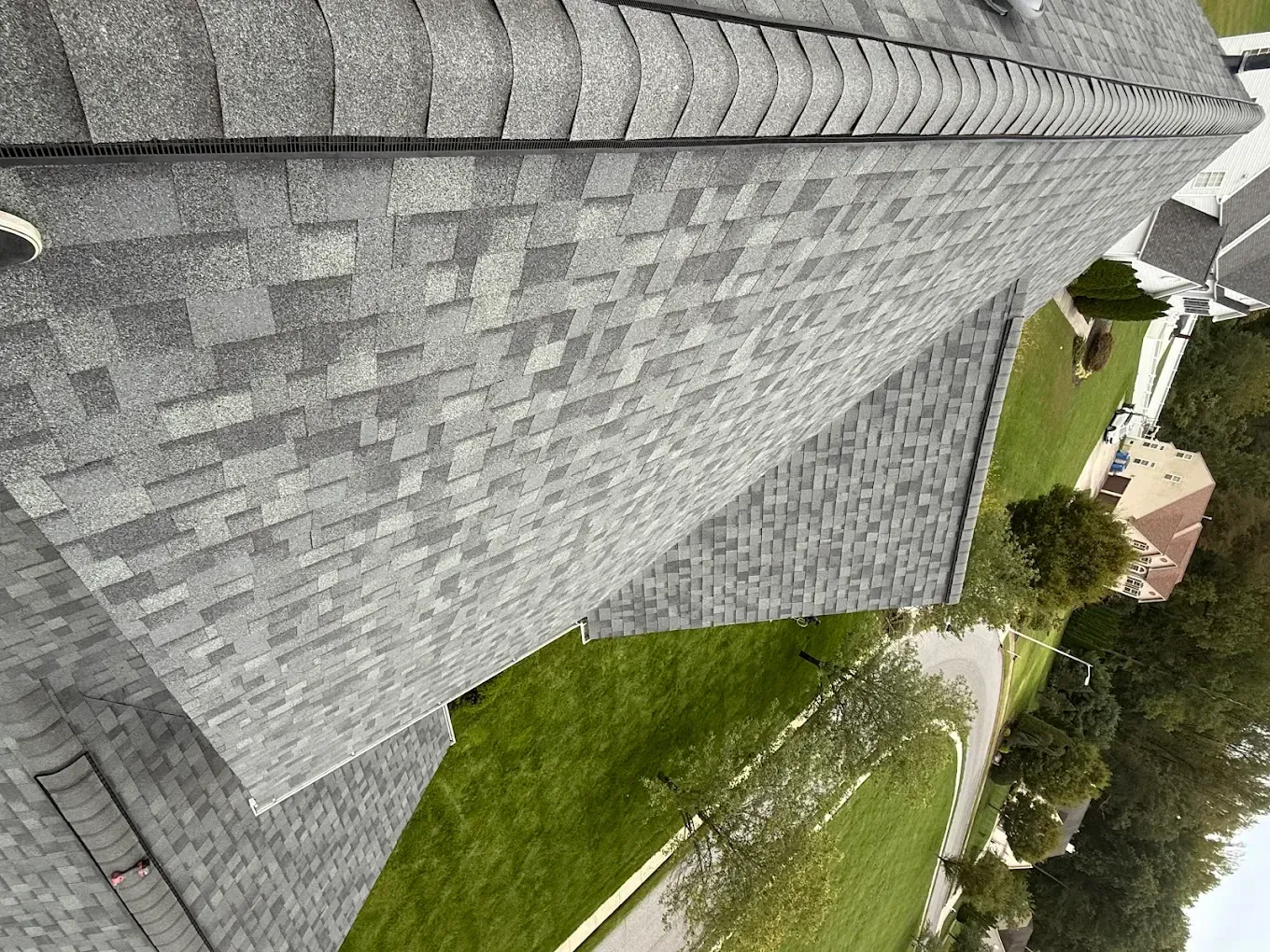 Tilted view of a gray shingled roof beside a curved green lawn and sidewalk near trees and houses