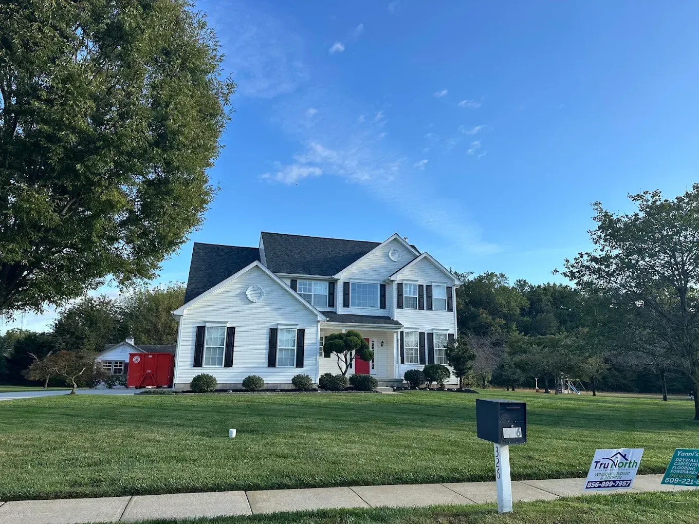 White two-story suburban house with a lawn, trees, and a mailbox under a blue sky