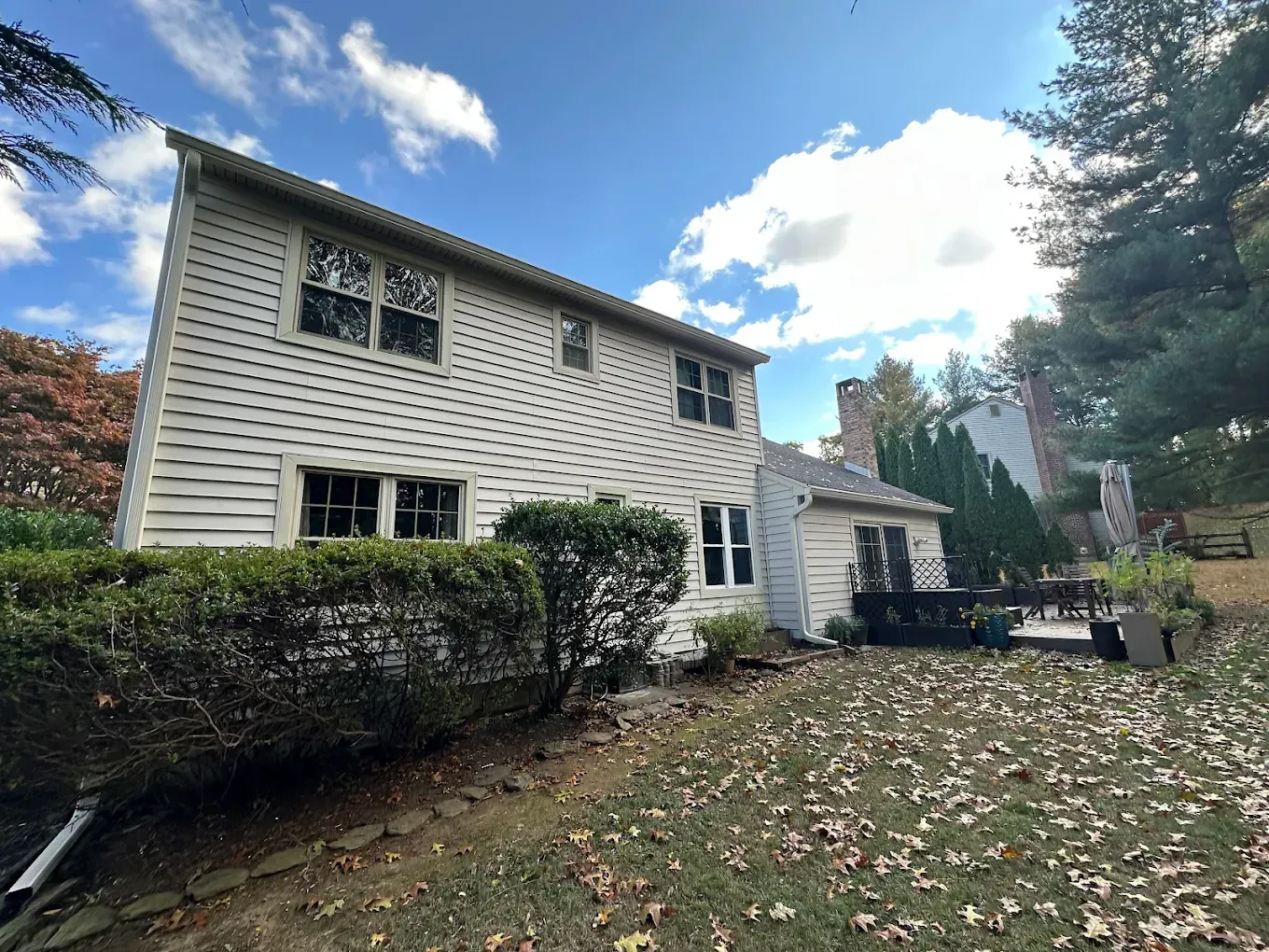Two-story white house with leaf-covered lawn, shrubs, and blue sky with clouds