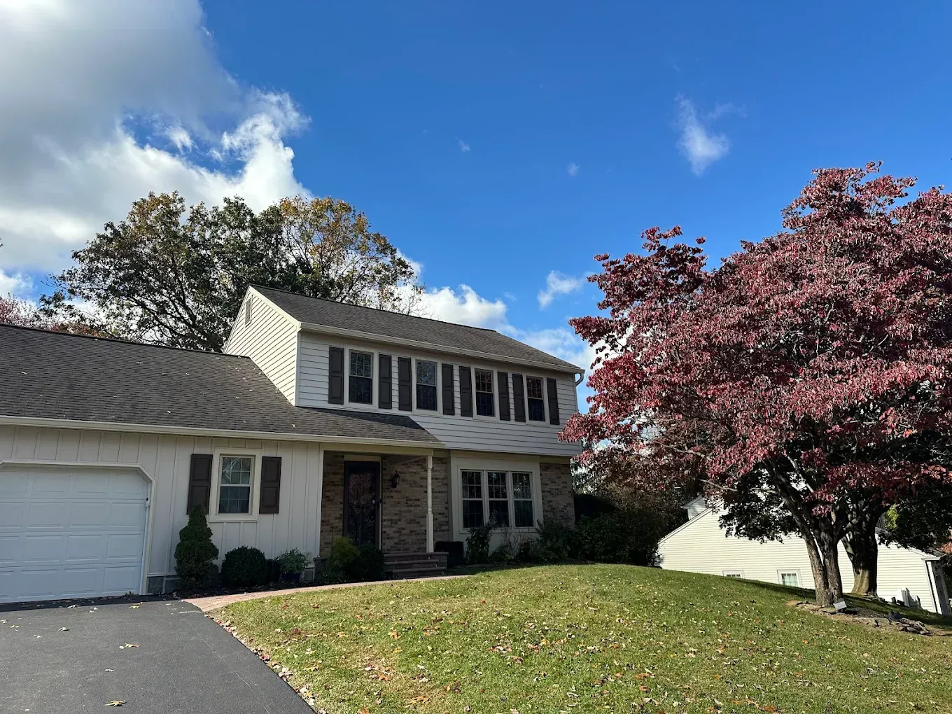 Two-story suburban house with white garage, stone entry, and a red-leaf tree under a blue sky