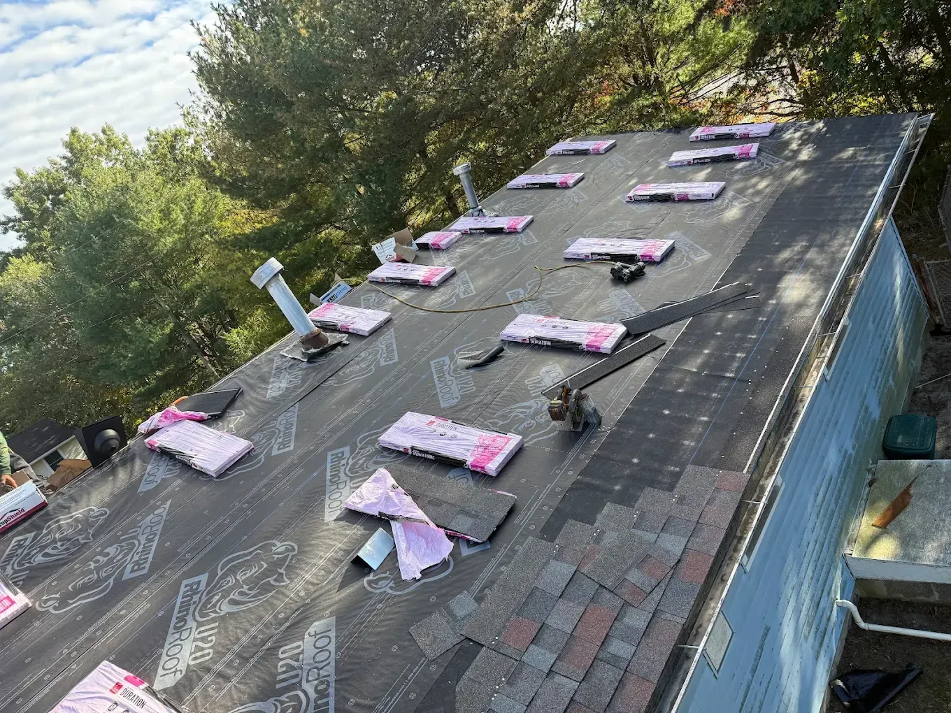 Roof with multiple pink-wrapped packages or panels arranged in rows on dark shingles, outdoors near trees