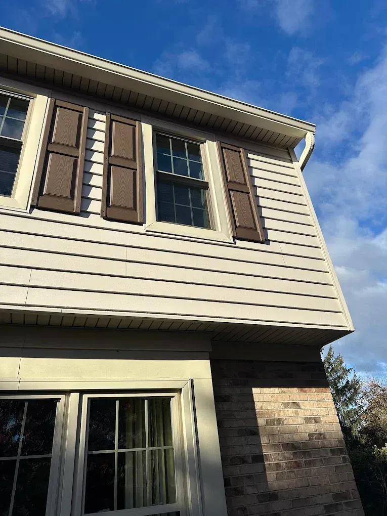 Two-story house exterior with tan siding, brown shutters, and a window under a blue sky