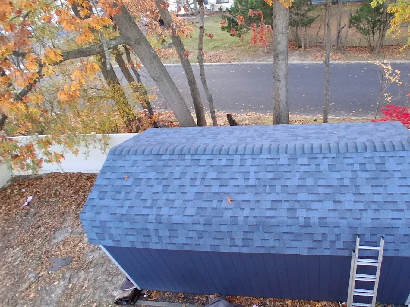 Aerial view of a blue-roofed shed beside a road, with a ladder leaning against it and autumn trees nearby