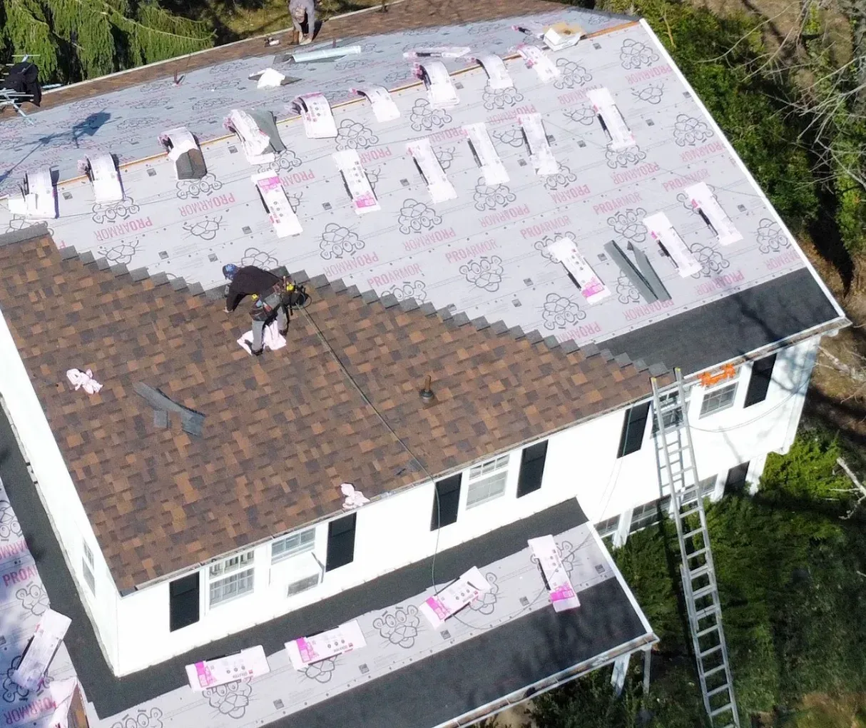 Roofers working on a house roof with brown shingles and pink underlayment sheets visible below.