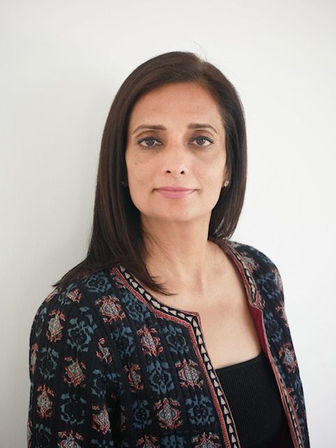 A woman wearing glasses and a striped shirt is standing in front of a white wall.
