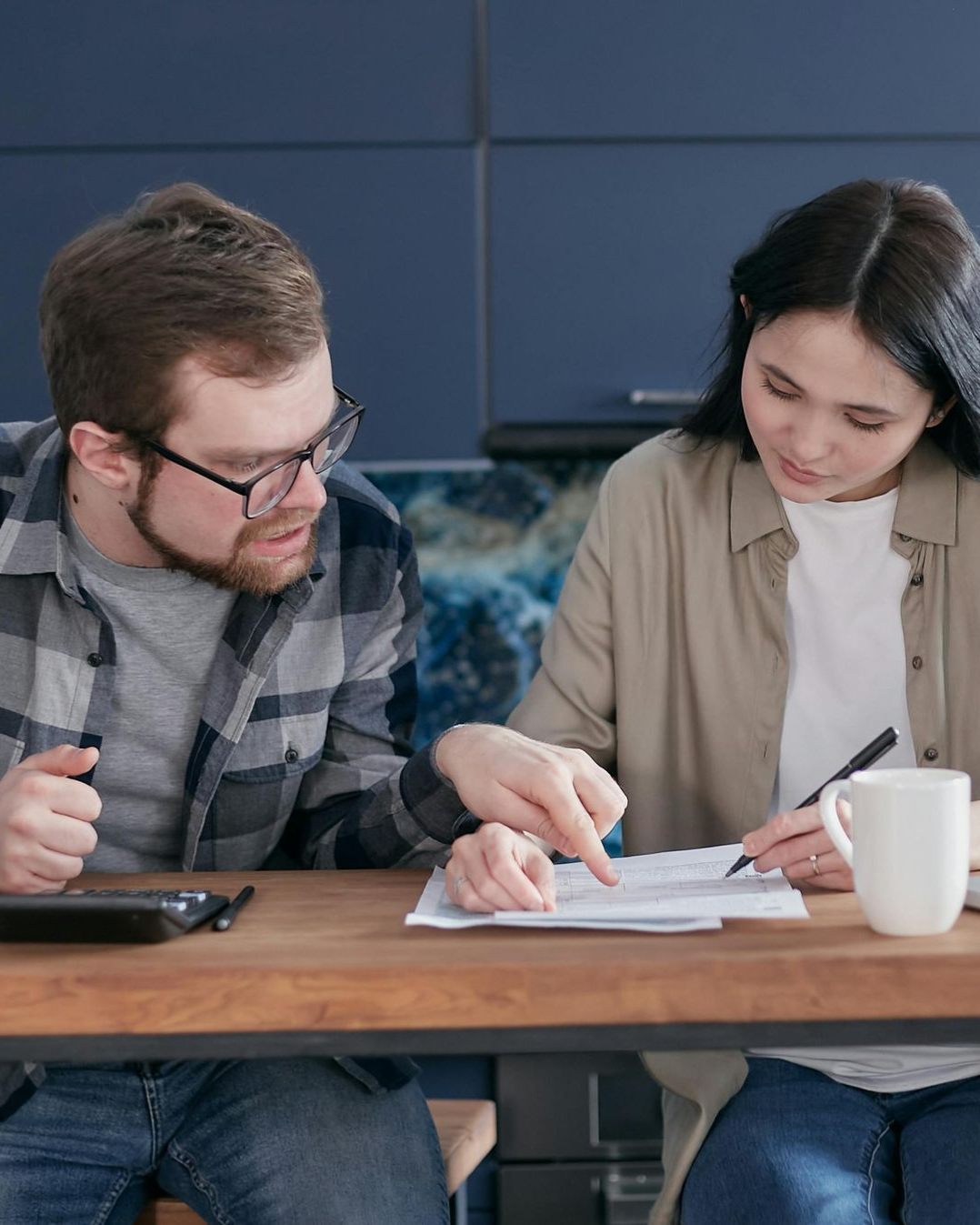 Two coworkers reviewing papers at a wooden table, with a laptop and coffee mug nearby.