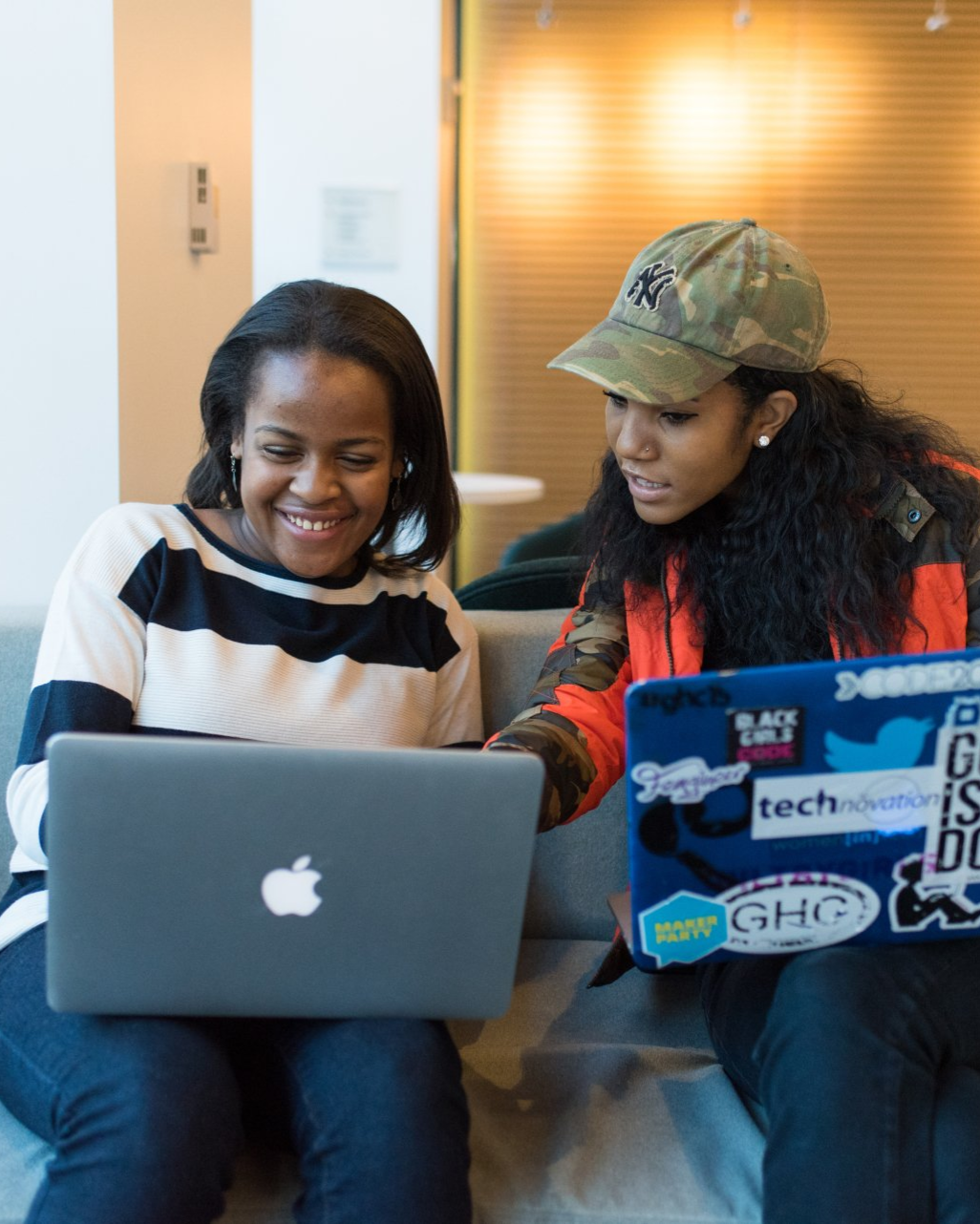 Students in a classroom. Some smiling and looking toward a person in front. Desks, window, bright lighting.