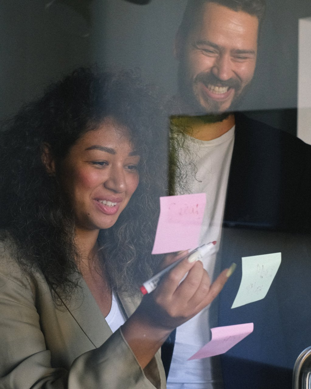 Two colleagues smiling and reading colorful sticky notes on a glass wall in an office