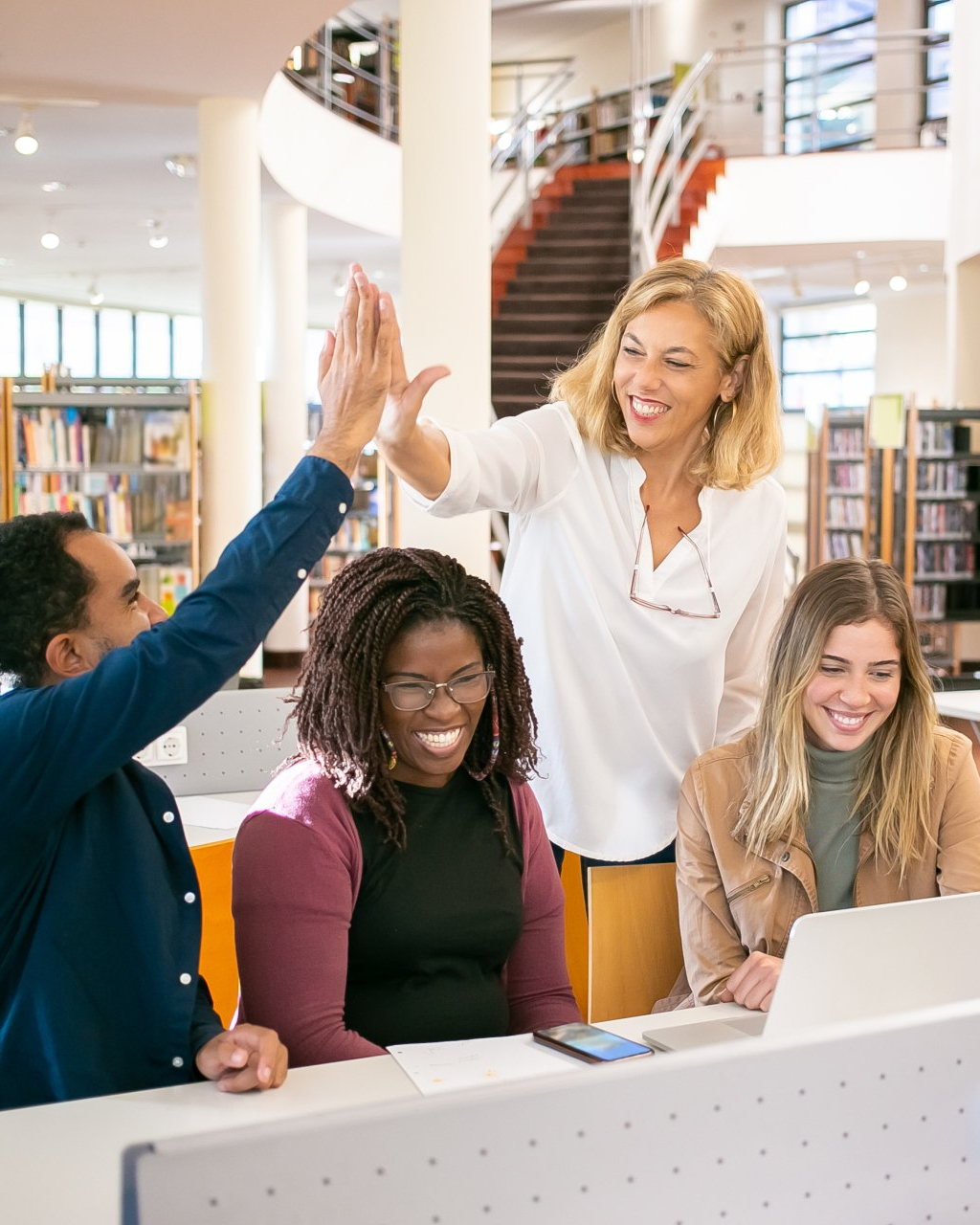 Group celebrating around a laptop in a bright library, with one person standing and raising a hand