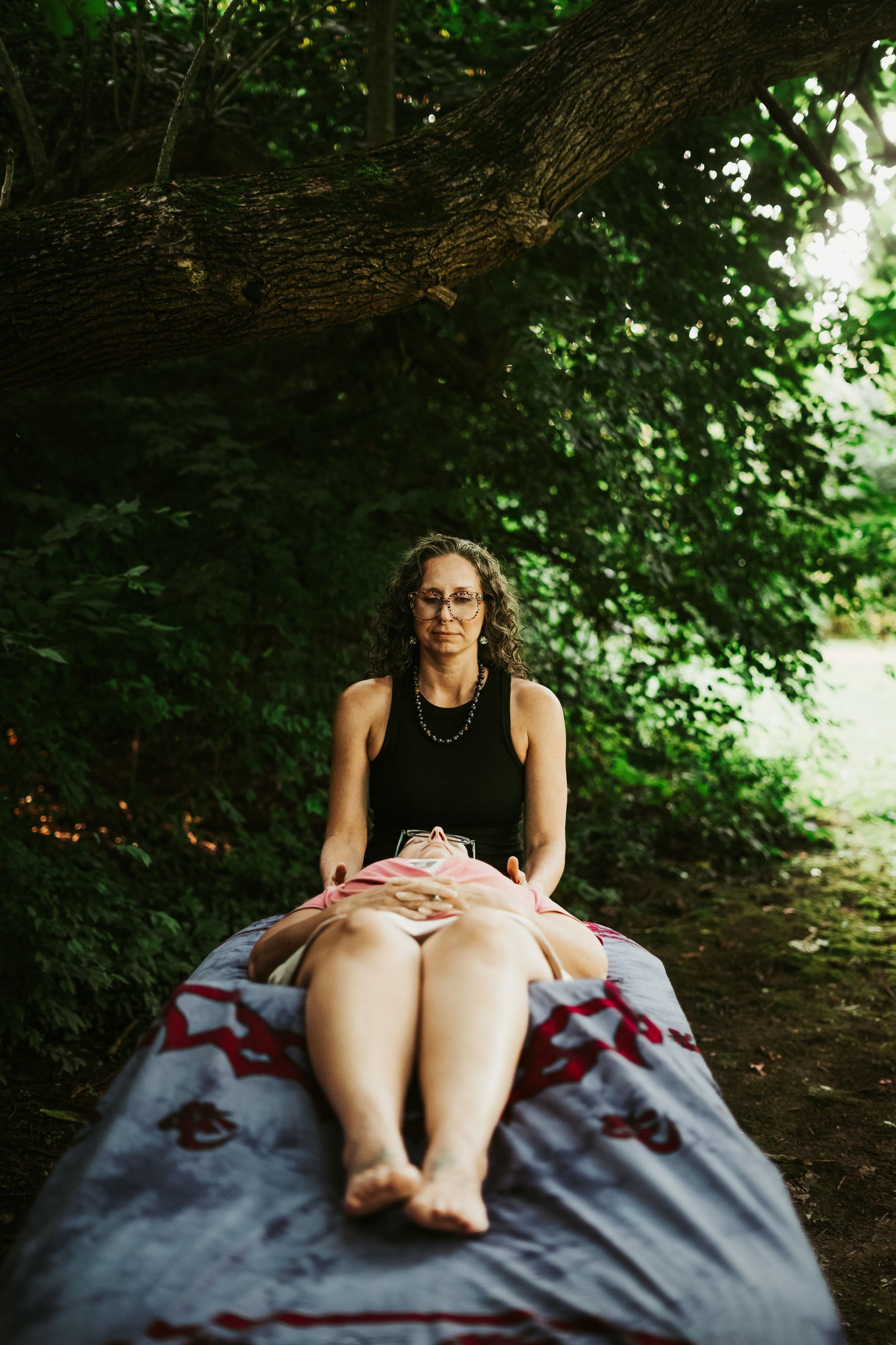 Woman giving massage outdoors on a gray patterned surface, beneath leafy trees.