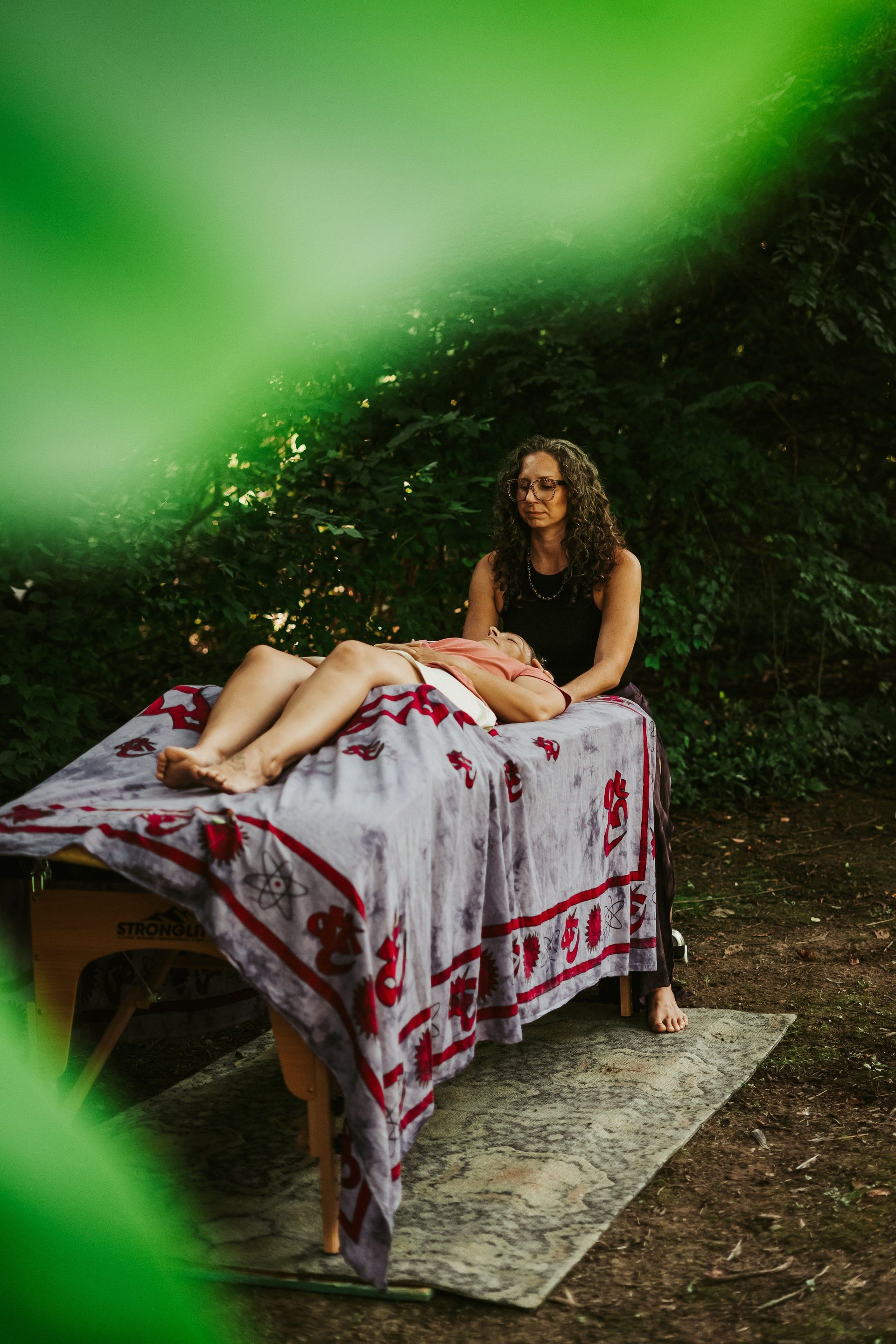 Woman giving massage outdoors on a massage table covered with a patterned blanket. Lush greenery surrounds them.