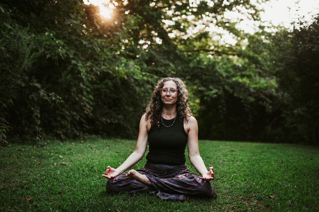 Woman meditating outdoors, cross-legged on grass, hands in mudra pose, smiling, surrounded by trees.