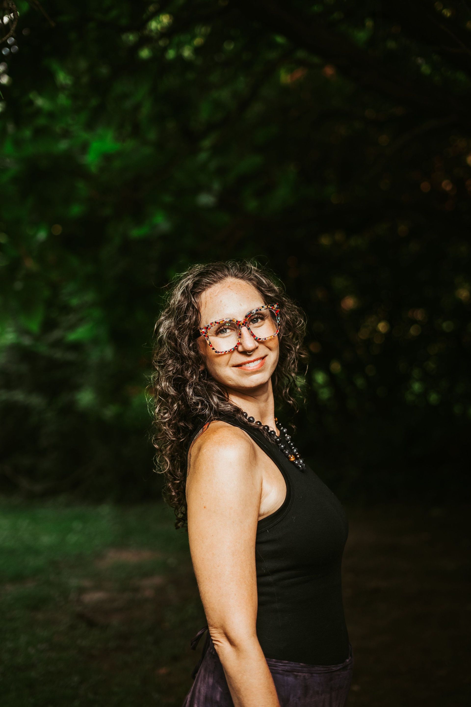 Woman with curly brown hair, glasses, and black top smiles outdoors near trees.