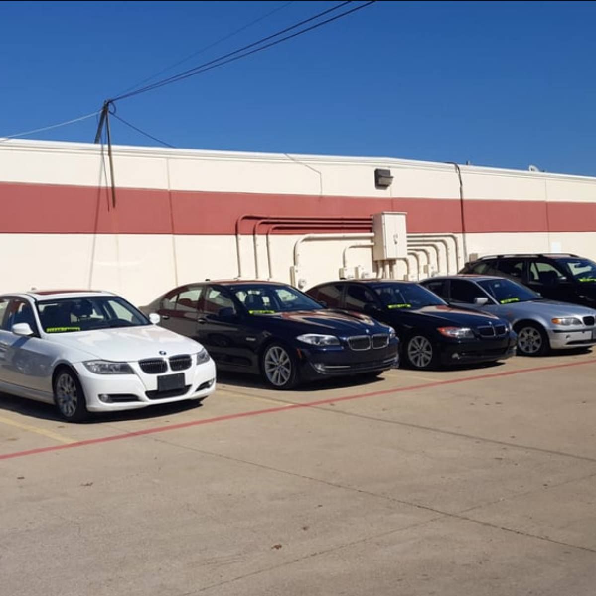 Several BMW sedans parked in front of a building with a red stripe. Sunny day.
