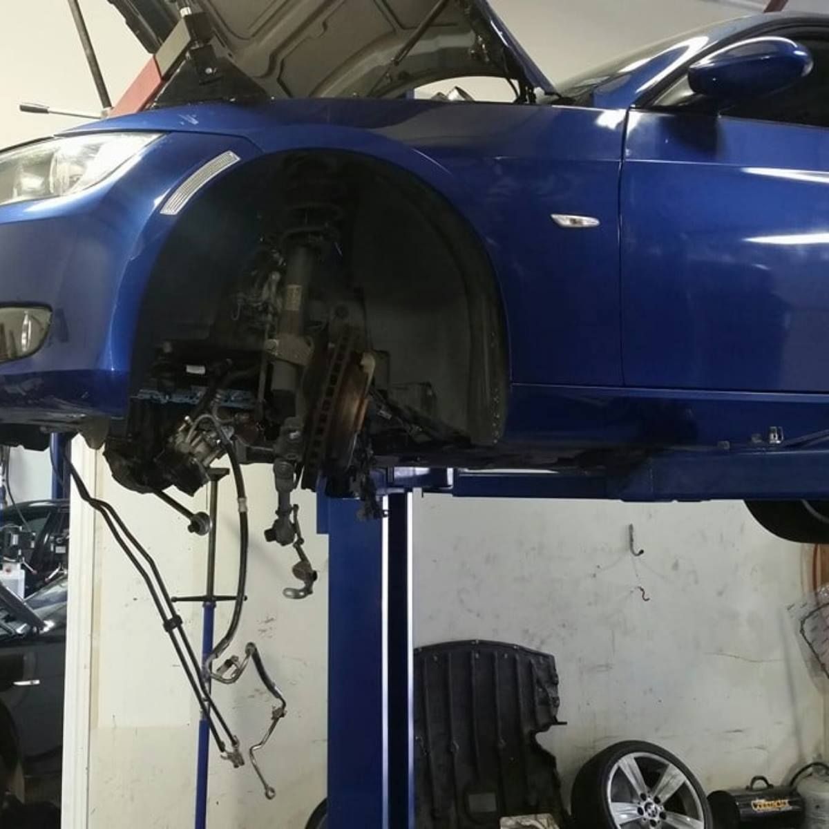 Blue car on a lift, front wheel assembly removed, in a repair shop.
